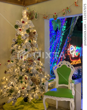 A snowy white Christmas tree decorated with lights and plush toys stands beside a green chair. The cozy indoor corner glows with warmth and holiday cheer. A snowy white Christmas tree decorated with lights and plush toys stands beside a green chair. The cozy indoor corner glows with warmth and holiday cheer. 132037075