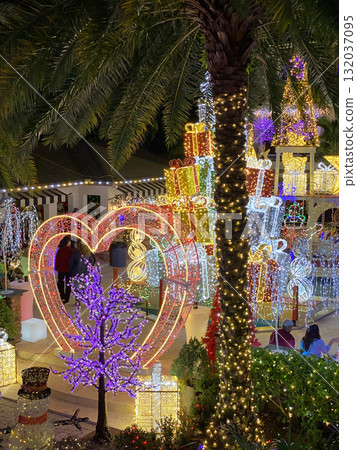 A glowing heart light stands beside palm trees surrounded by bright Christmas gifts. The colorful decorations create a warm tropical holiday mood. A glowing heart light stands beside palm trees surrounded by bright Christmas gifts. The colorful decorations create a warm tropical holiday mood. 132037095