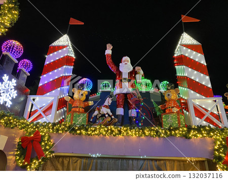 Large Santa Claus figure stands between tall red towers decorated with garlands, ribbons, and colorful lights. The festive outdoor scene captures the joyful Christmas spirit. Large Santa Claus figure stands between tall red towers decorated with garlands, ribbons, and colorful lights. The festive outdoor scene captures the joyful Christmas spirit. 132037116