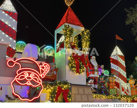 A large Santa decoration stands on a lighted tower with red ribbons and Christmas ornaments. The nighttime holiday scene glows with color and festive warmth. 132037120