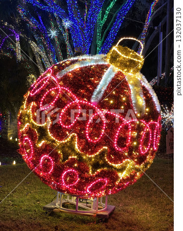 Bright red Christmas ornament with glowing lights stands in a park surrounded by trees decorated with blue and green lights. Festive scene shines with holiday spirit. 132037130