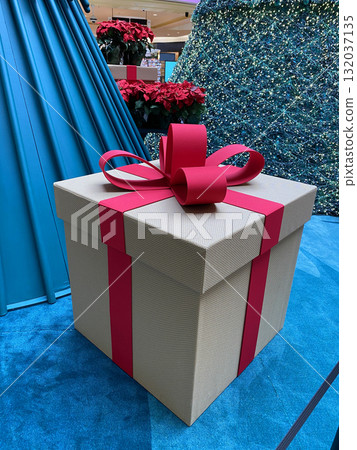 Close-up of a big decorative gift box with a red ribbon and bow inside a Christmas mall installation. The festive present stands near sparkling trees and bright holiday decor. Close-up of a big decorative gift box with a red ribbon and bow inside a Christmas mall installation. The festive present stands near sparkling trees and bright holiday decor. 132037135