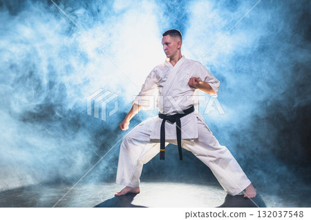 Strong man fighter in white kimono for karate martial arts with black belt preparing for fight training in black background. Man training moves punches kicks practicing shadow fight ready for sparring 132037548