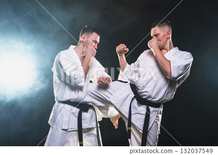 Martial arts masters on dark background. Strong man fighters in white kimono for karate martial arts fighting training in black gym. Karate fighters training punches kicks practicing fight sparring Martial arts masters on dark background. Strong man fighters in white kimono for karate martial arts fighting training in black gym. Karate fighters training punches kicks practicing fight sparring 132037549