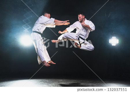 Martial arts masters on dark background. Strong man fighters in white kimono for karate martial arts fighting training in black gym. Karate fighters training punches kicks practicing fight sparring Martial arts masters on dark background. Strong man fighters in white kimono for karate martial arts fighting training in black gym. Karate fighters training punches kicks practicing fight sparring 132037551