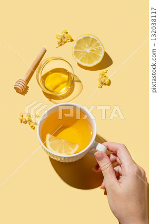 Hand holding cup of herbal tea with lemon and honey on a yellow background with dry flowers 132038117