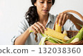 Woman peeling fresh corn on the cob from a wicker basket. Close-up of hands preparing a raw vegetable. Healthy eating and farm-to-table harvest concept 132038360