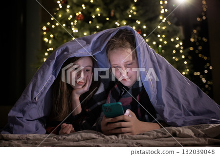 Two girls under a blanket looking at a smartphone near a Christmas tree with festive lights 132039048