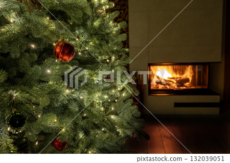 Beautiful close-up of a decorated Christmas tree with glowing lights and red ornaments beside a cozy fireplace, symbolizing warmth and festive comfort. 132039051