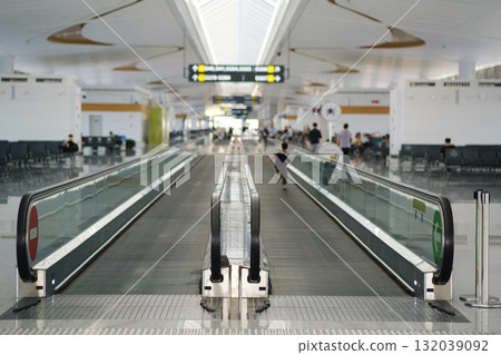 Wide and modern airport terminal with moving walkway and people walking, showcasing a bright and spacious environment during daytime 132039092