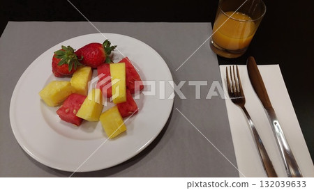 Healthy breakfast with fresh pineapple, watermelon, and strawberries on a white plate, served with orange juice and cutlery on a minimalist table setting, top view 132039633