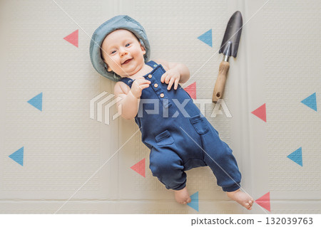 Adorable baby dressed as a little gardener with a small garden shovel nearby. Concept of gardening, nature connection, and playful early childhood imagination 132039763