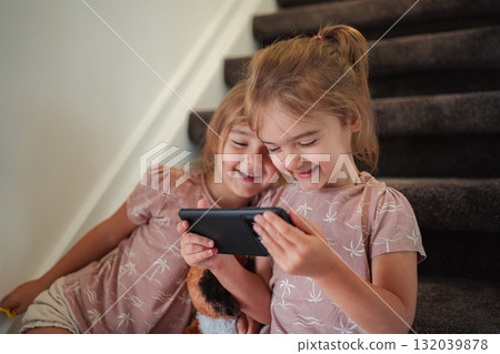 Two little sisters laughing and playing with a smartphone sitting on stairs at home Two little sisters laughing and playing with a smartphone sitting on stairs at home 132039878