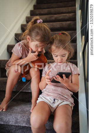 Two preschool girls playing with smartphone and teddy bear on stairs Two preschool girls playing with smartphone and teddy bear on stairs 132039879