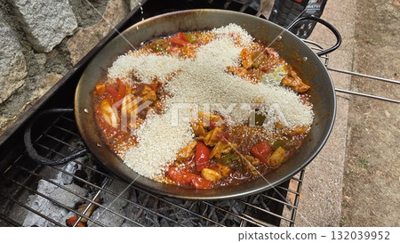 Traditional spanish paella cooking outdoors over open flame with rich ingredients. Rice in the shape of a cross in paella. Typical cuisine of Spain. Stage of cooking paella. Ingredients for paella Traditional spanish paella cooking outdoors over open flame with rich ingredients. Rice in the shape of a cross in paella. Typical cuisine of Spain. Stage of cooking paella. Ingredients for paella 132039952