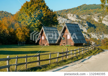 Closer view of two triangular wooden houses behind a rustic fence with mountains and trees in the background. Warm autumn scene, showcasing rural architecture and peaceful countryside atmosphere. Closer view of two triangular wooden houses behind a rustic fence with mountains and trees in the background. Warm autumn scene, showcasing rural architecture and peaceful countryside atmosphere. 132040395