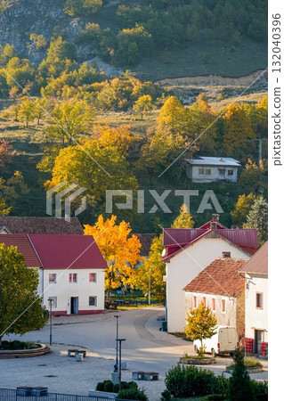 Scenic view of a pair of houses with red roofs situated in front of rolling hills covered with colorful autumn trees. Warm fall colors create a peaceful and picturesque rural atmosphere. Ideal for 132040396