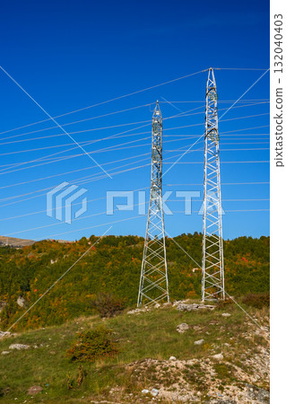 Electric power pole standing on a grassy hill with a scenic landscape in the background. The structure carries electricity across the area, surrounded by natural hills and trees. Suitable for energy Electric power pole standing on a grassy hill with a scenic landscape in the background. The structure carries electricity across the area, surrounded by natural hills and trees. Suitable for energy 132040403