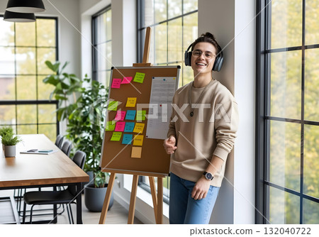 Neurodivergent man wearing headphone smiling beside corkboard with colorful sticky notes in modern meeting room with large windows and indoor plants 132040722