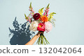 A hand holds a beautiful, elegant bouquet of fresh flowers against a bright, light blue background. 132042064