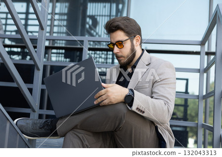 Man in suit and sunglasses sitting on steps, focused on laptop screen. Man in suit and sunglasses sitting on steps, focused on laptop screen. 132043313