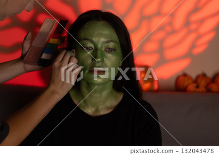 A person is applying green makeup to another person's face to create a Halloween A person is applying green makeup to another person's face to create a Halloween 132043478