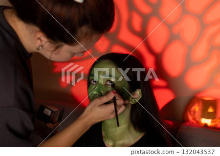 Woman is applying green face paint to a woman's face to create a scary Halloween 132043537