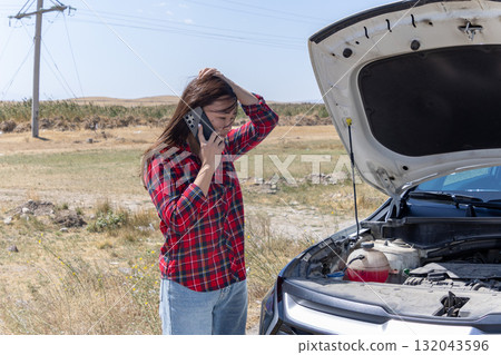 A distressed woman is on her phone next to a broken-down car with the hood open 132043596