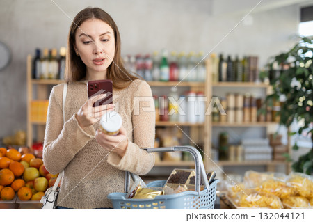 Young woman customer with shopping basket buying food in grocery store, reading ingredients on jar of conserve asparagus and scan QR code Young woman customer with shopping basket buying food in grocery store, reading ingredients on jar of conserve asparagus and scan QR code 132044121