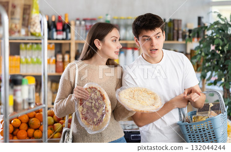 Happy young wife and husband choose large and fresh pizza in supermarket in department of semi-finished products 132044244