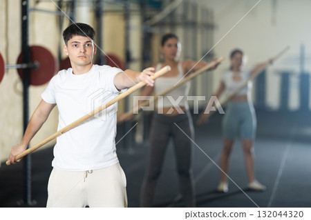 Young guy, together with other athletes, does a warm-up using a long stick in gym 132044320