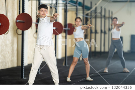 Young guy, together with other athletes, does a warm-up using a long stick in gym Young guy, together with other athletes, does a warm-up using a long stick in gym 132044336