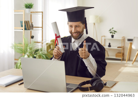 Man in graduate gown sitting at desk with diploma showing thumb up sign during online video call. Man in graduate gown sitting at desk with diploma showing thumb up sign during online video call. 132044963