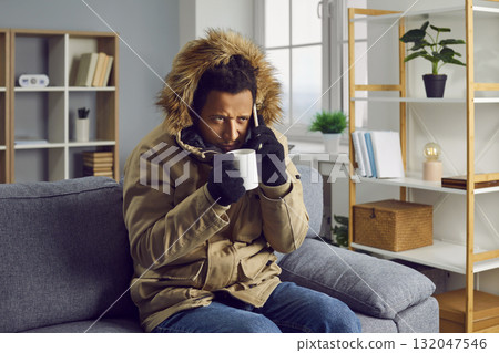 Anxious young man is talking on the phone and drinking hot drink, sitting at home in knitted gloves and warm jacket with hood, freezing from cold, solves problem remotely. 132047546