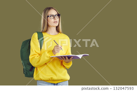 Serious blondy student girl in glasses with backpack makes notes in copybook on khaki background. Serious blondy student girl in glasses with backpack makes notes in copybook on khaki background. 132047593
