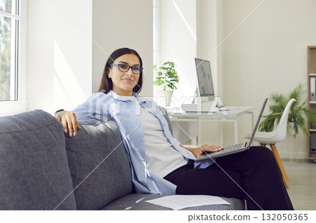 Portrait of young businesswoman sitting on sofa with laptop and looking at camera Portrait of young businesswoman sitting on sofa with laptop and looking at camera 132050365