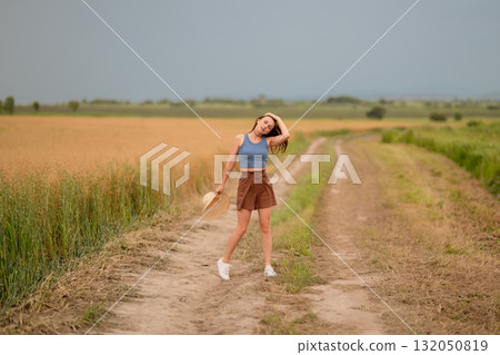 Young woman enjoying a sunny day in a wheat field while posing along a dirt road 132050819