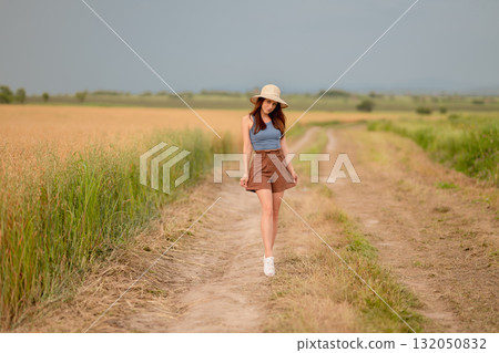 Young woman walks along a rural path in warm sunlight surrounded by golden fields 132050832