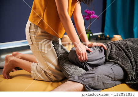 Masseuse making thai yoga massage. Cropped view of woman therapist doing traditional massage treatment for male patient. Client lying on mat, meditating, enjoying hand massage in calm atmosphere. Masseuse making thai yoga massage. Cropped view of woman therapist doing traditional massage treatment for male patient. Client lying on mat, meditating, enjoying hand massage in calm atmosphere. 132051033