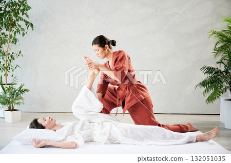 Masseuse making thai yoga massage. Woman therapist doing traditional massage treatment, stretching female patient's body. Client lying on mat, enjoying hand massage in calm atmosphere. Masseuse making thai yoga massage. Woman therapist doing traditional massage treatment, stretching female patient's body. Client lying on mat, enjoying hand massage in calm atmosphere. 132051035