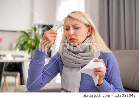 Blonde middle aged woman lying on a sofa covered with blanket feeling unwell and checking fever with thermometer while sick at home 132051042