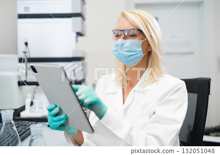 Female scientist in lab coat and mask using tablet to analyze data in laboratory. 132051048