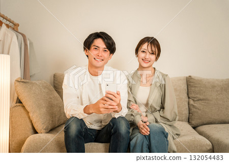 一對年輕的亞洲夫婦在家裡的客廳沙發上一邊看智慧型手機一邊聊天 132054483