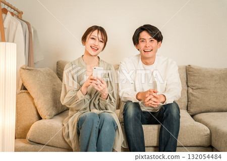 一對年輕的亞洲夫婦在家裡的客廳沙發上一邊看智慧型手機一邊聊天 一對年輕的亞洲夫婦在家裡的客廳沙發上一邊看智慧型手機一邊聊天 132054484