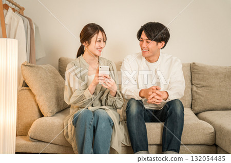 一對年輕的亞洲夫婦在家裡的客廳沙發上一邊看智慧型手機一邊聊天 一對年輕的亞洲夫婦在家裡的客廳沙發上一邊看智慧型手機一邊聊天 132054485