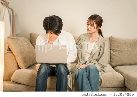 A wife looks anxiously at her husband working on a computer on the sofa in the living room (overtime, taking work home, hidden overtime) 132054899