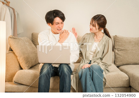 A wife looks anxiously at her husband working on a computer on the sofa in the living room (overtime, taking work home, hidden overtime) 132054900