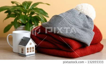 A knitted hat in a warm shade rests beside a small model house, symbolizing comfort and coziness in winter 132055333