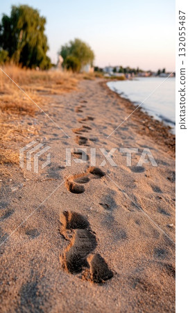 Footprints lead across a sandy beach as the sun sets, creating a serene atmosphere with waves softly touching the shore 132055487