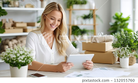 A happy woman is looking at a tablet, enjoying her online shopping experience at a neat workspace filled with greenery and packages 132055760